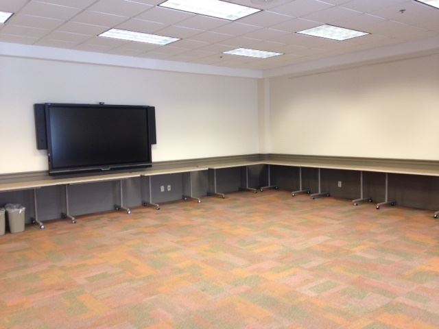 Carpeted meeting room with tables pushed against the wall. Large monitor for use on left wall.