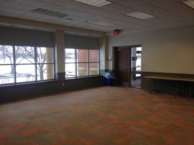 Large carpeted meeting room. Two sets of shaded windows shown next to door into library entry